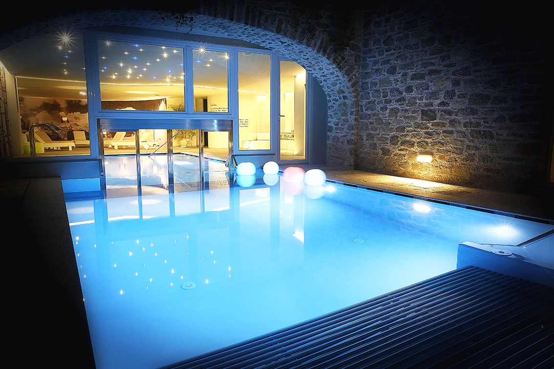 A swimming pool with a glass enclosure, a stone wall, and a well-lit interior.