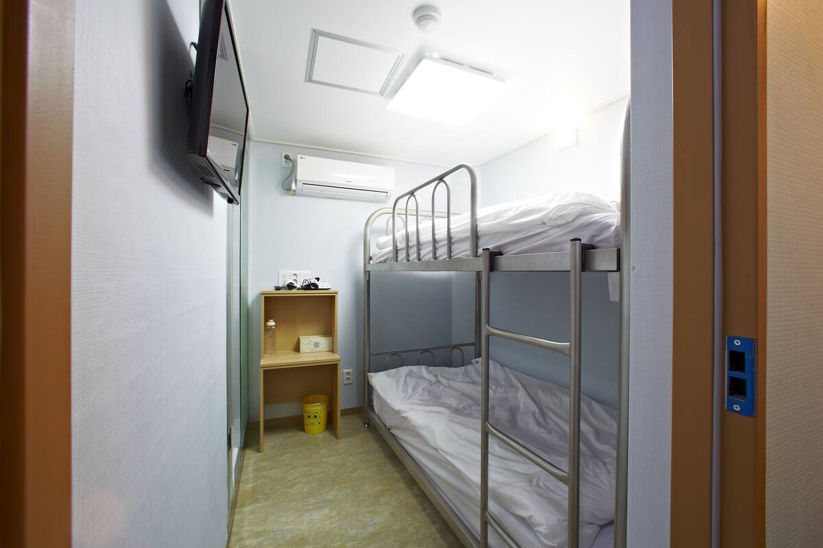 Habitació petita i ben il·luminada amb lliteres, una prateleira de fusta i un safataire.