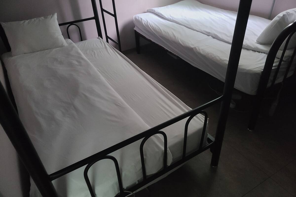 Un llit individual amb estructura metàl·lica i roba de llit blanca en una habitació amb sòl de rajola.