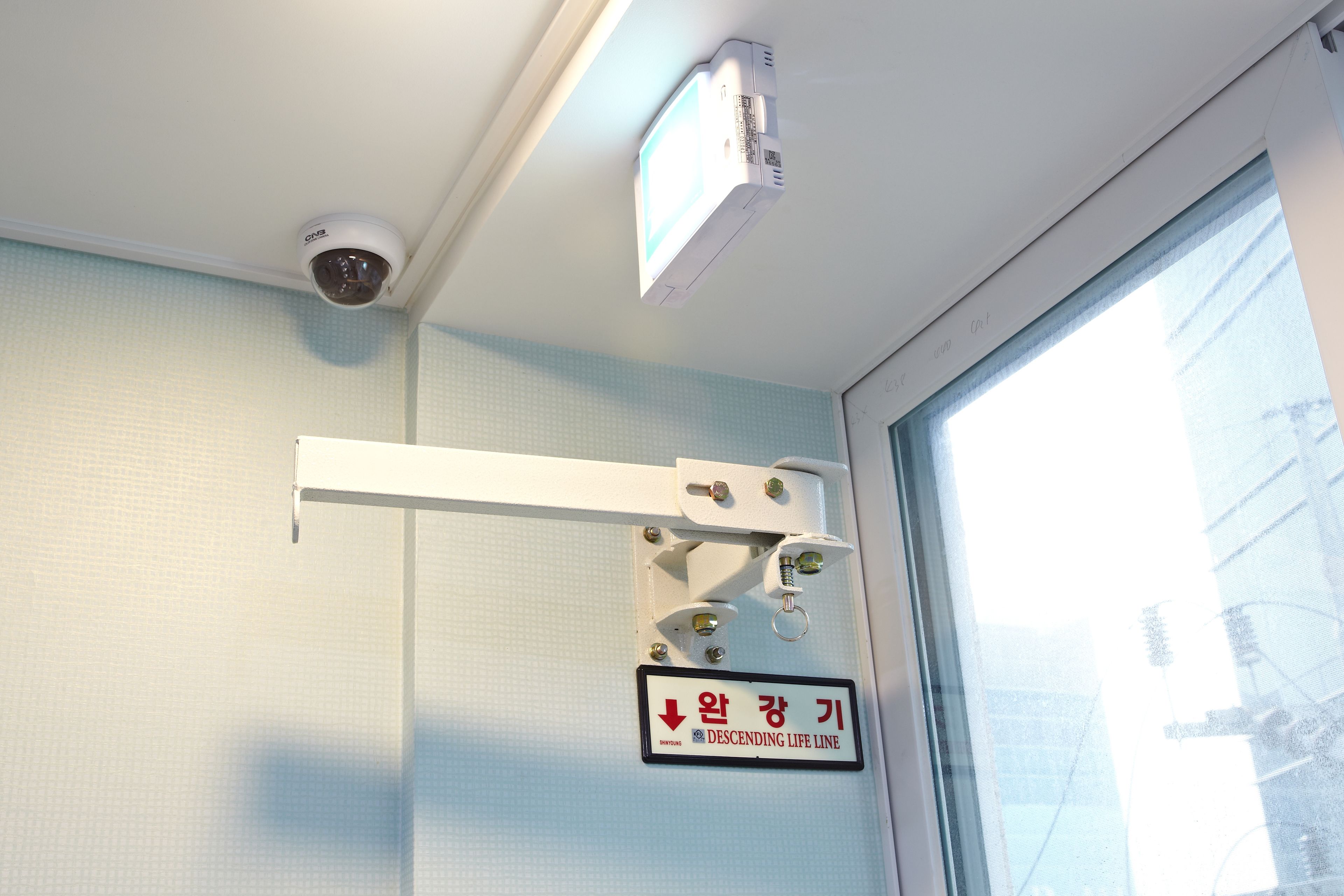 A room with a white ceiling, a security camera, a window with a view of the outside, and a sign with Korean text.