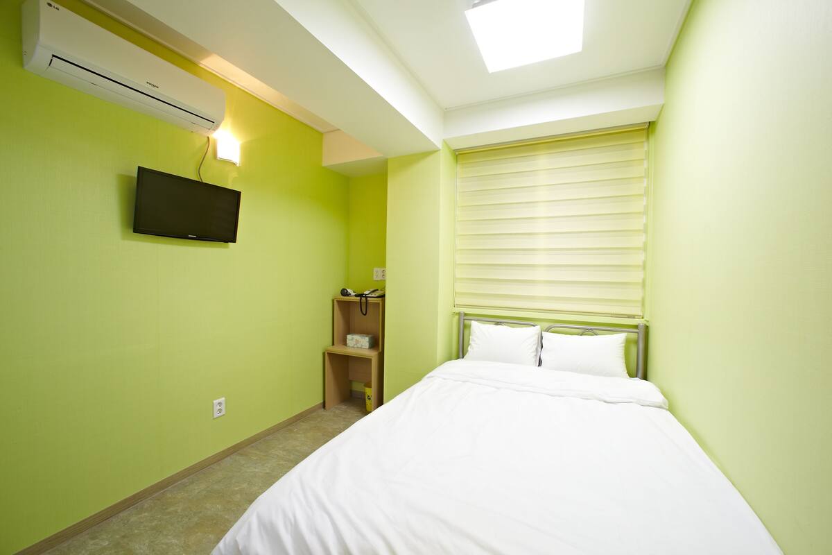 Habitació d'hotel amb un llit, un televisor penjat a la paret, aire condicionat, una taula de nit amb una làmpada i una finestra amb persianes.