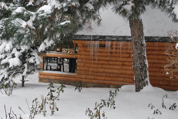 A log cabin with a wooden deck and a stone foundation, surrounded by snow-covered ground and trees. - Elki-Palki (Sovinion)