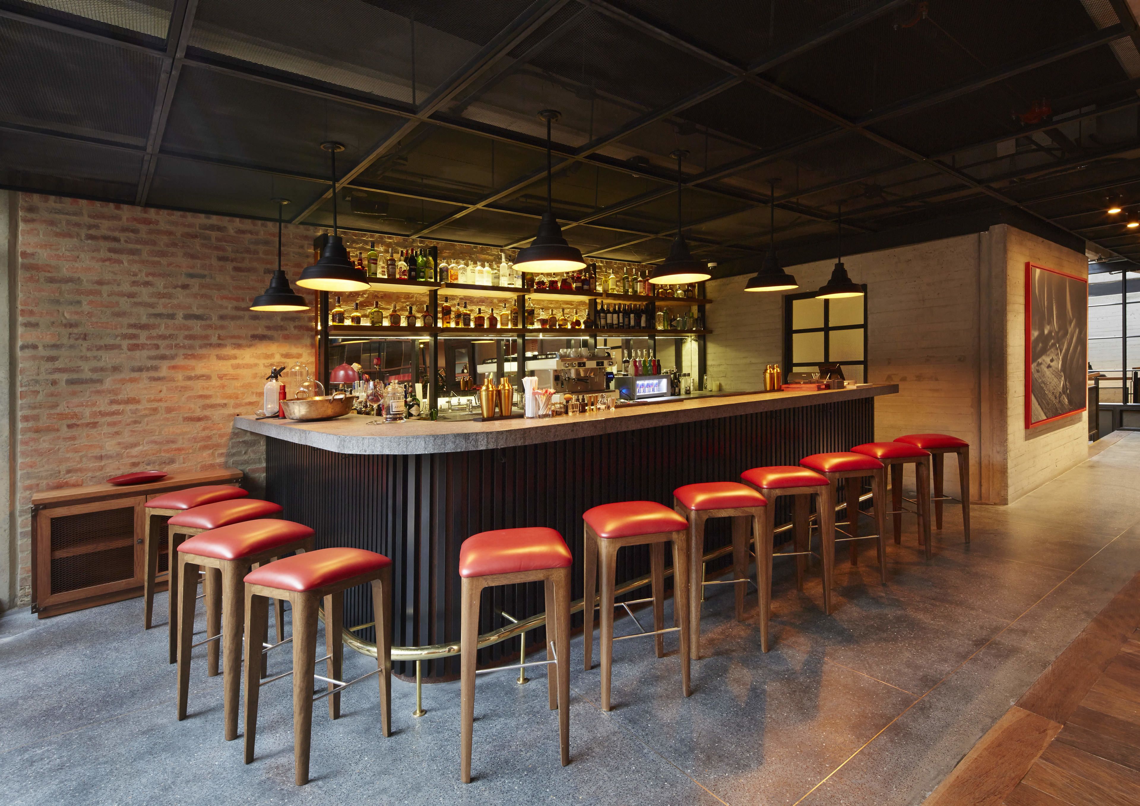 Zona de bar con pared de ladrillo, taburetes de madera y barra con varias botellas y vasos.