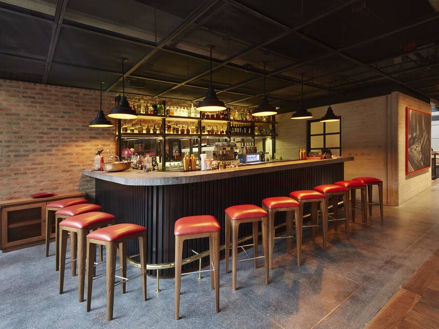 Zona de bar con pared de ladrillo, taburetes de madera y barra con varias botellas y vasos.