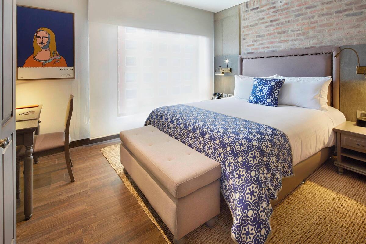A modern hotel room with a large bed, a wooden bench, a desk, and a framed artwork on the wall.