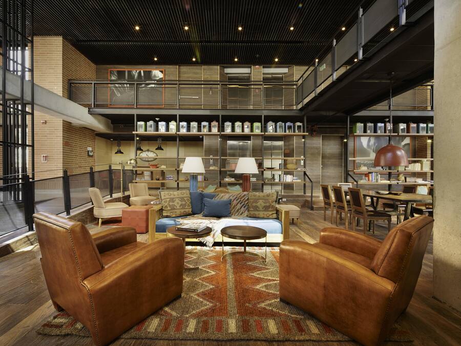 Uma área de estar ampla com poltronas de couro, um sofá e uma mesa de jantar. Há prateleiras com diversos objetos e um mezanino com assentos adicionais.