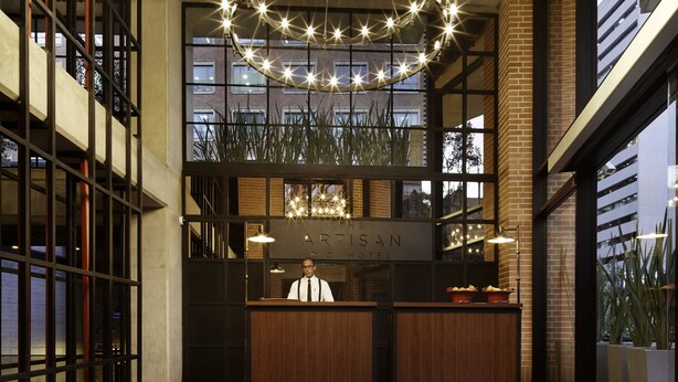 Een hotel lobby met een moderne kroonluchter, een houten receptiebalie en grote ramen met uitzicht op een gebouw.