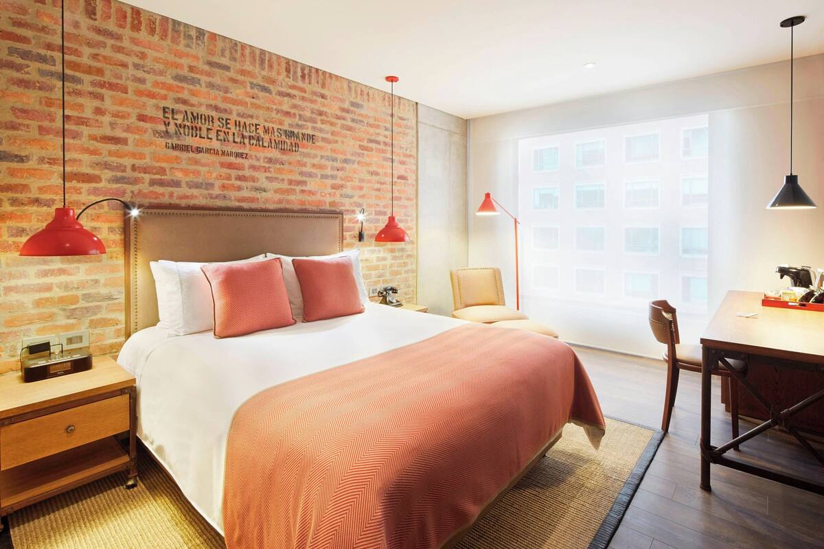 A hotel room with a brick wall, a large bed with a headboard, two red pendant lights, a wooden nightstand, a desk with a chair, and a large window.