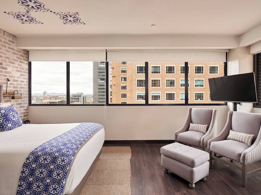 Habitación de hotel con una cama grande, dos sillones, un televisor de pantalla plana y vistas a un paisaje urbano a través de amplios ventanales.