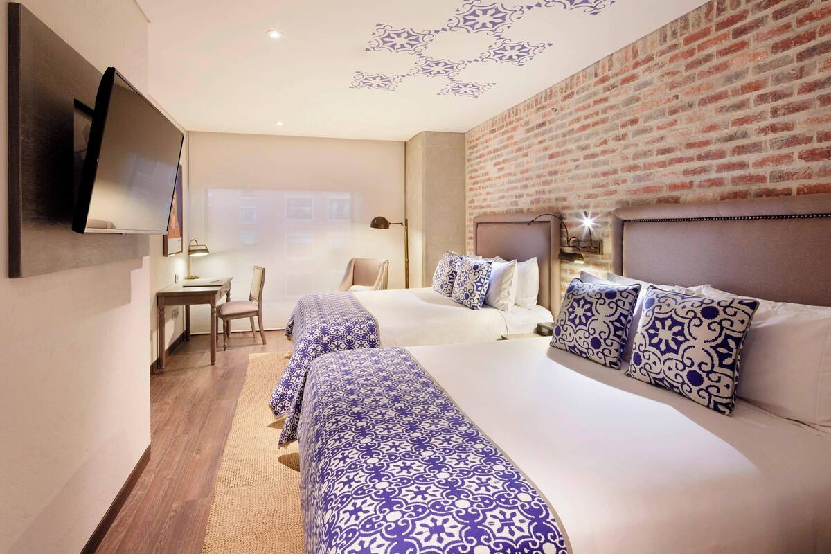 A hotel room with two beds, a flat-screen TV mounted on the wall, a desk with a chair, and a brick-patterned wall.