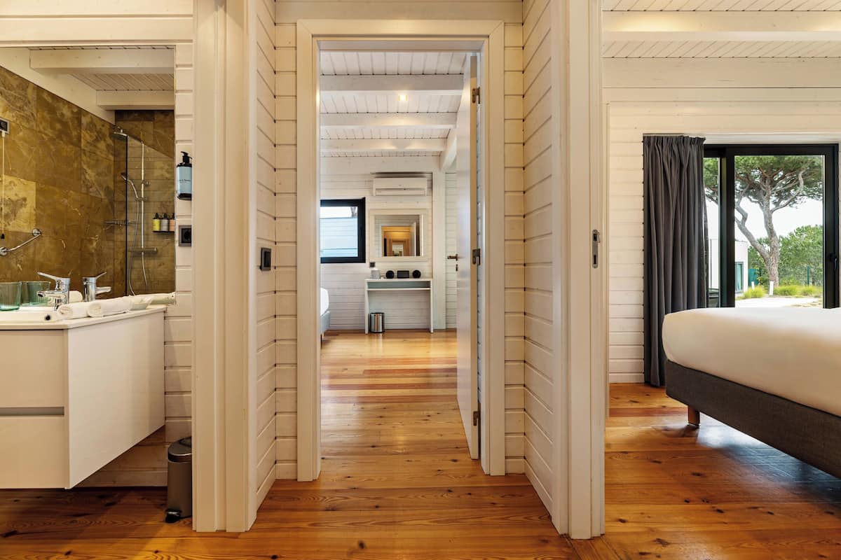 Habitación de hotel moderna con piso de madera, una cama grande, baño con espejo y lavamanos, y vista al exterior.