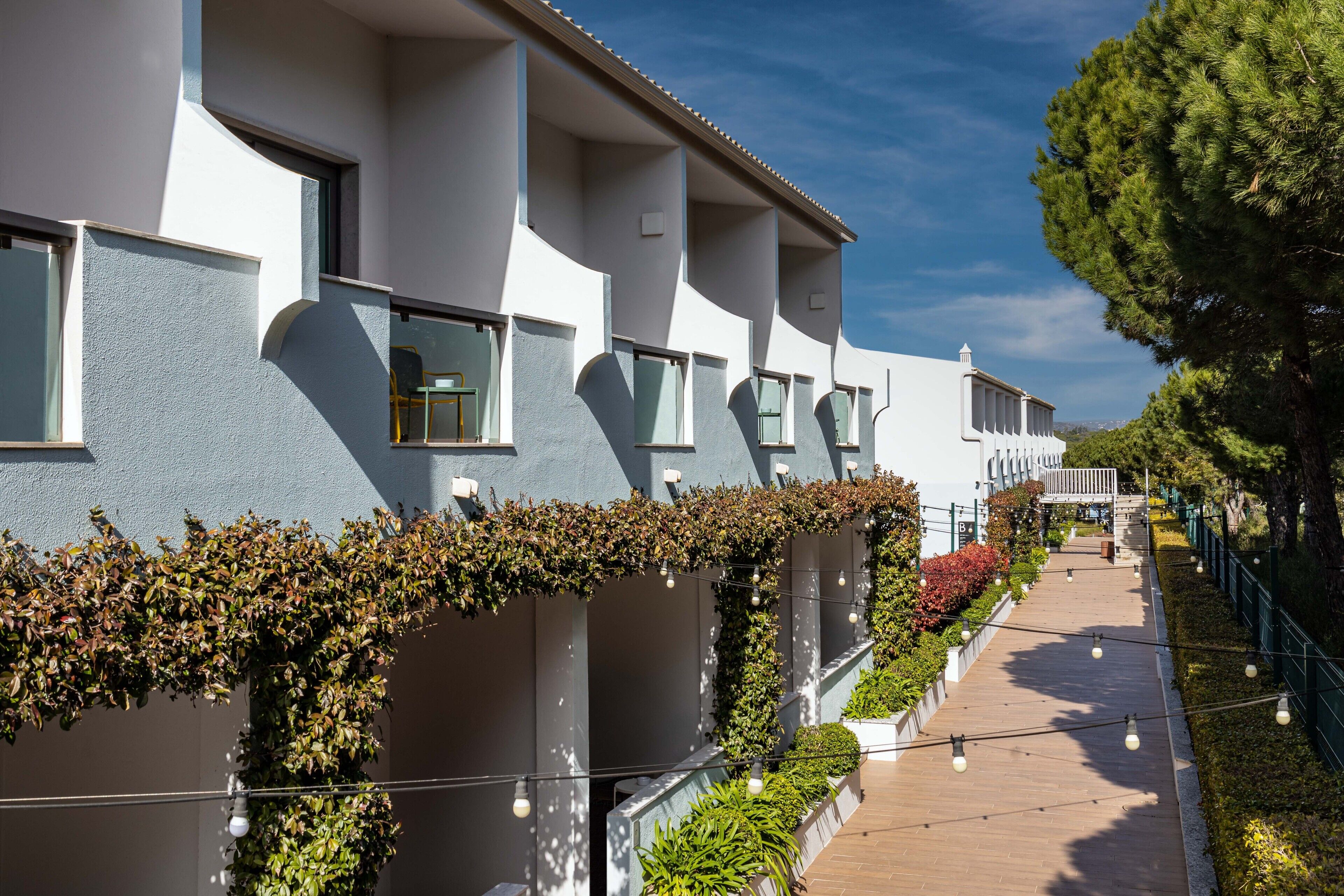 Un hotel moderno con una fila de balcones, un paseo de madera y vegetación a un lado.