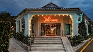 L’entrée d’un hôtel, avec un panneau néon indiquant « Bienvenue », un escalier menant à l’étage supérieur et un jardin bien entretenu.