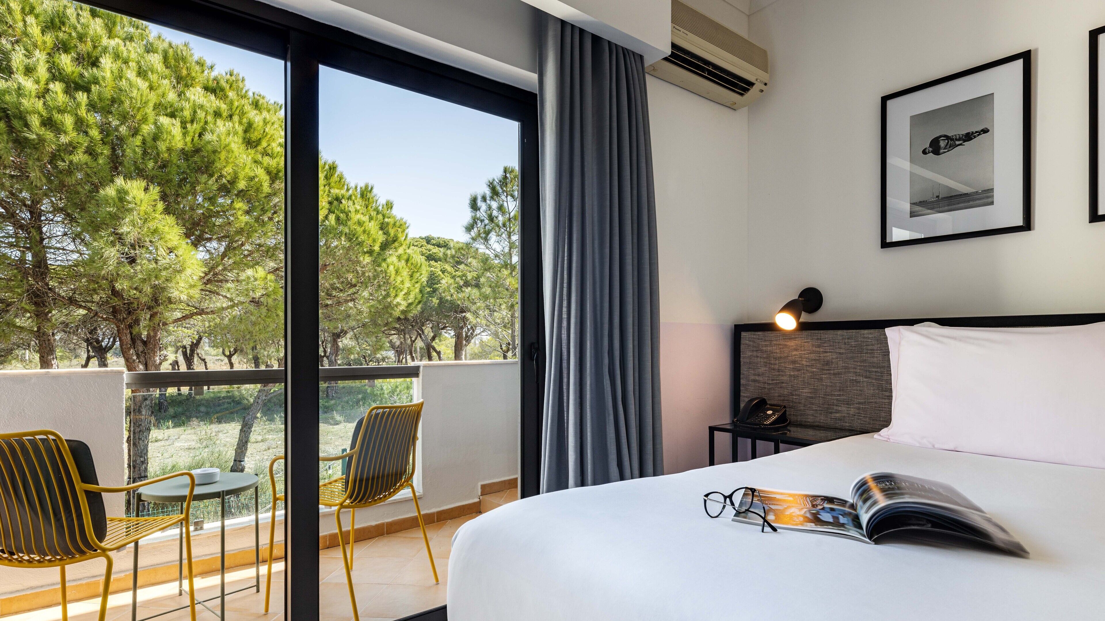 A hotel room with a bed, a bedside table with a lamp, a chair, and a view of trees through large windows.