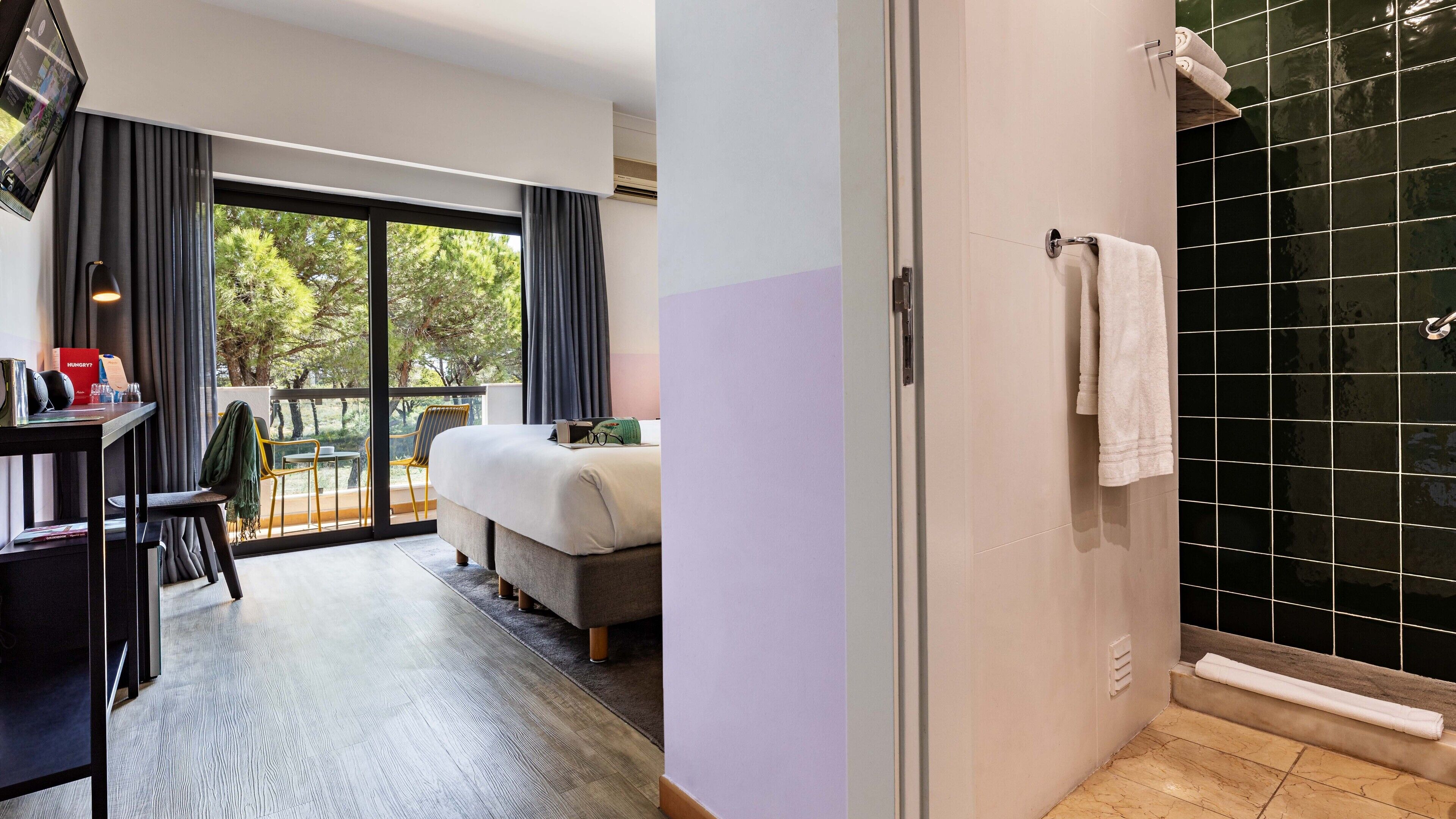 Habitación de hotel con cama, escritorio con computadora, una silla y vista a árboles a través de amplias ventanas.