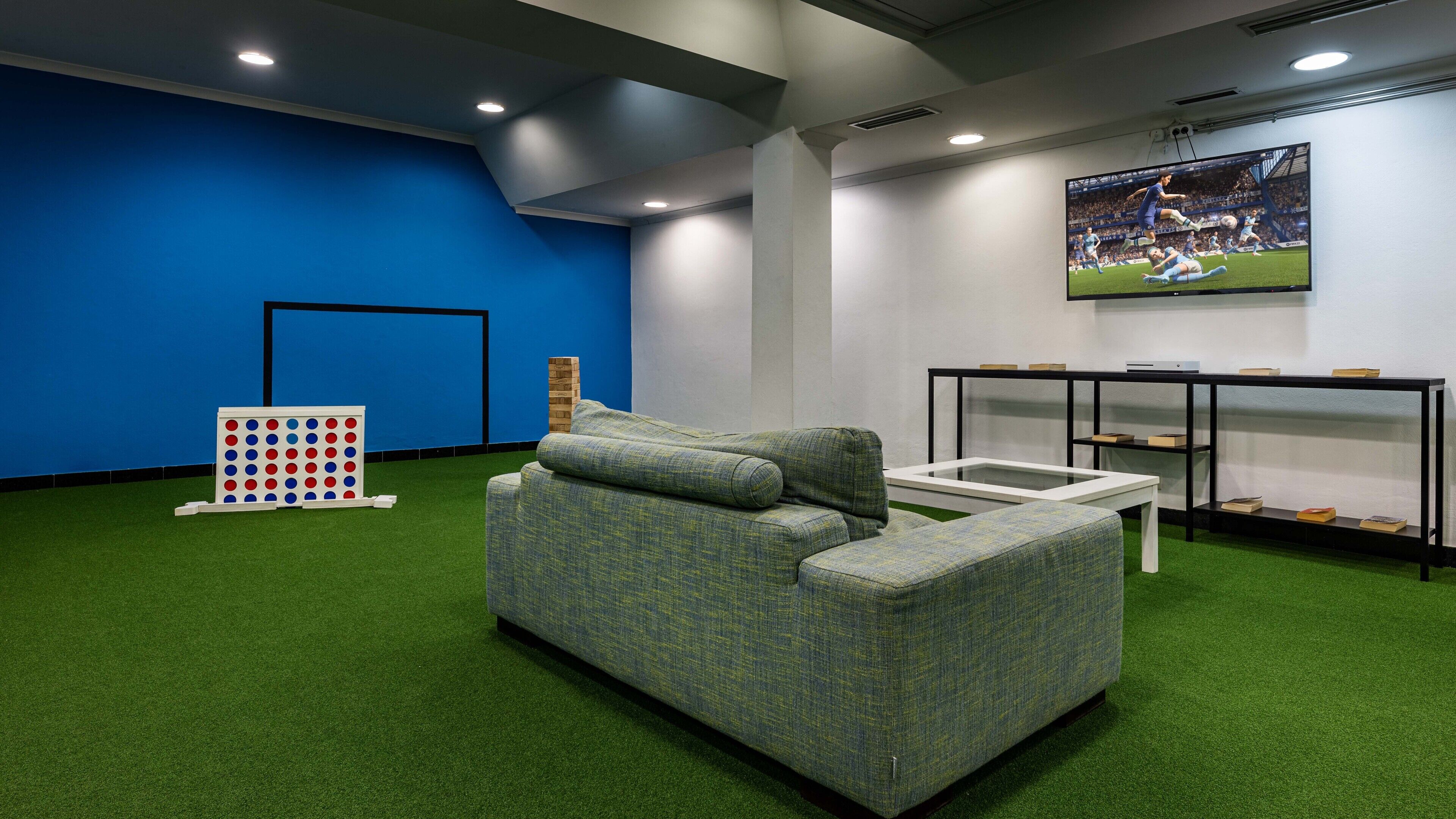 A room with a blue wall, a sectional sofa, a game board, and a television displaying a soccer match.