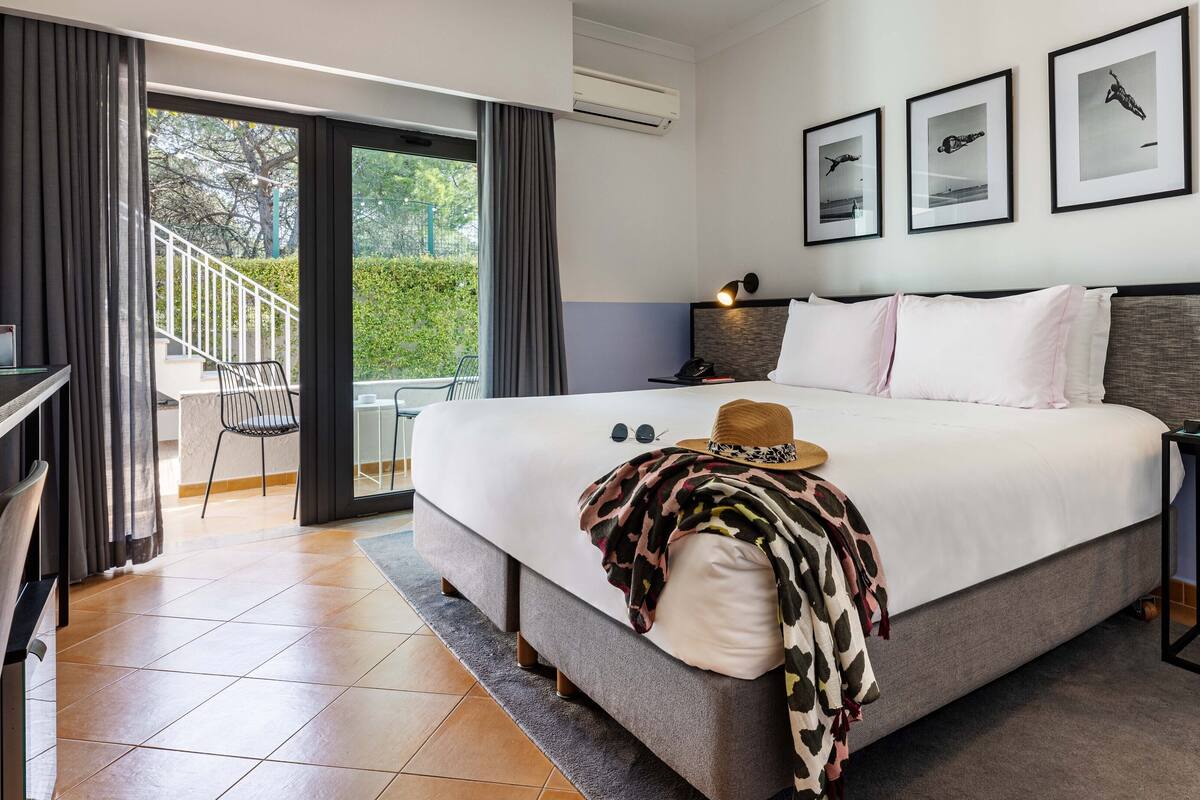 Habitación de hotel con cama, escritorio, silla y balcón con vistas a la vegetación.