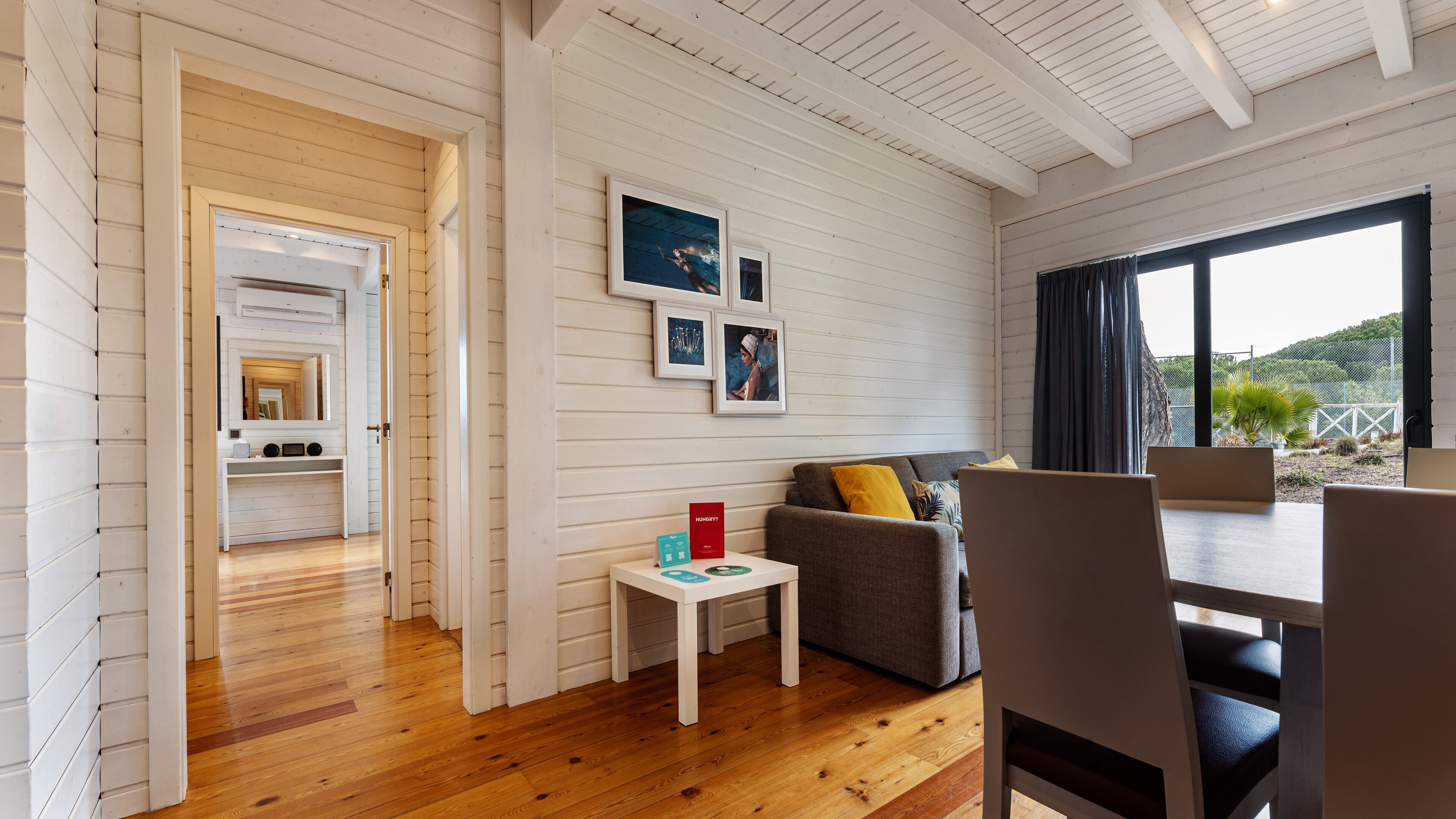 Un salon moderne avec un plancher en bois, un canapé, une table basse et un coin repas avec des chaises. On peut apercevoir la cuisine à travers une porte ouverte.