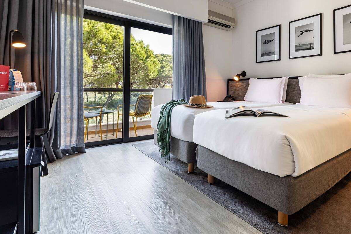 Una habitación de hotel moderna con una cama grande, un escritorio con silla, un comedor con sillas y un balcón con vistas a árboles.