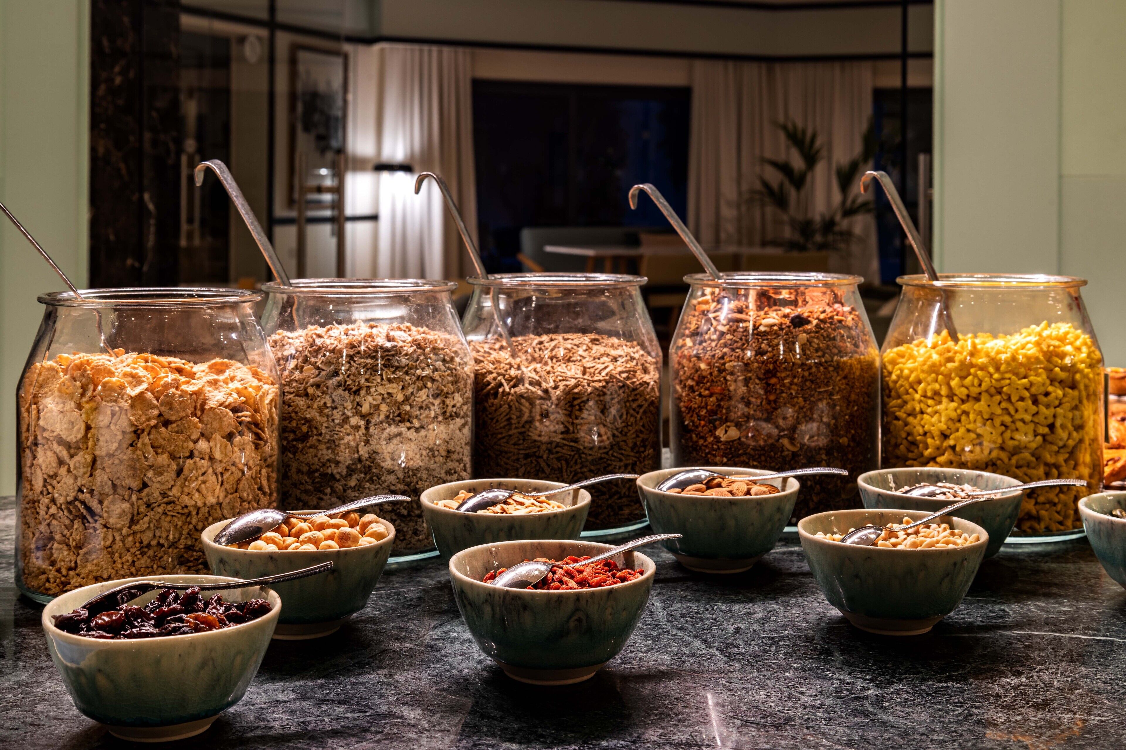 A buffet with various food items in bowls and jars, including cereals, nuts, and dried fruits.