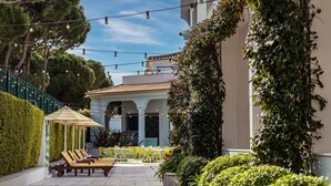 Un espace extérieur aménagé avec des chaises jaunes et des parasols à rayures, entouré de verdure et bordé d’un bâtiment dont le toit est recouvert de tuiles.