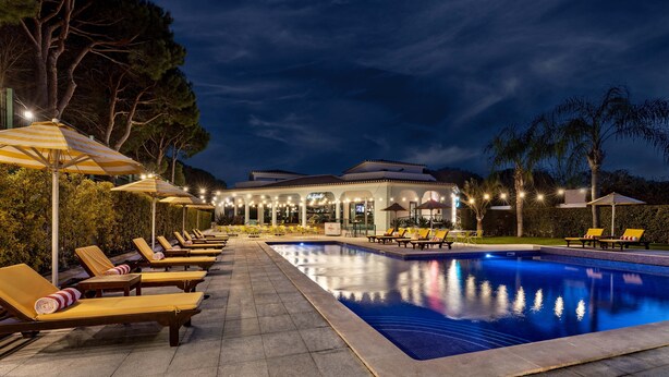 Un espace aménagé au bord de la piscine, avec des chaises longues et des parasols, une piscine et un bâtiment illuminé.