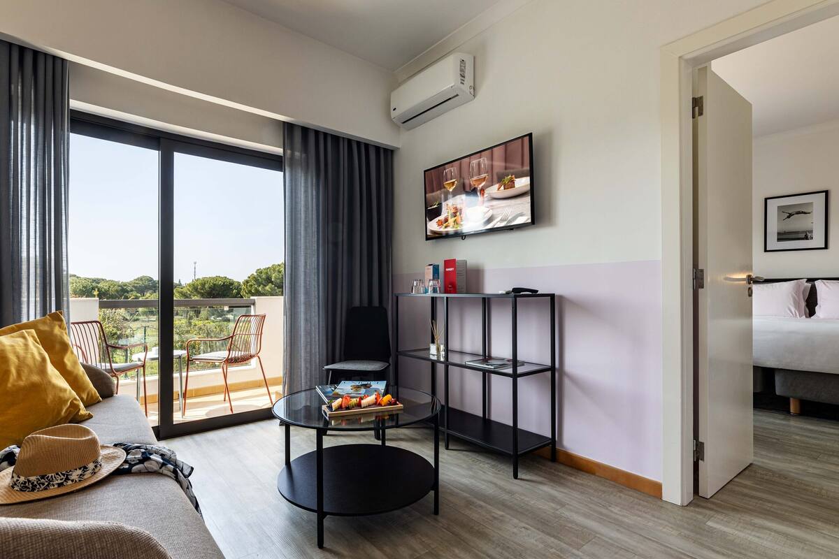 Una habitación de hotel moderna con balcón, un sofá con cojines amarillos, una mesita redonda con una bandeja de frutas, una estantería de metal negro con libros y un jarrón, y una cama con ropa blanca.