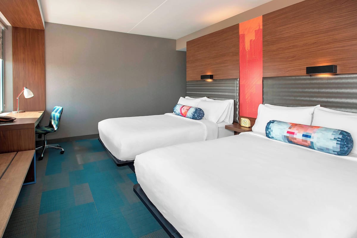 Une chambre d’hôtel avec deux lits, un bureau, une chaise et une fresque représentant un paysage urbain sur le mur.