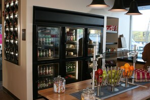 A bar area with a refrigerated display case, a wooden counter with a fruit bowl, and a view of the street outside. - Aloft by Marriott Buffalo Airport (Buffalo)