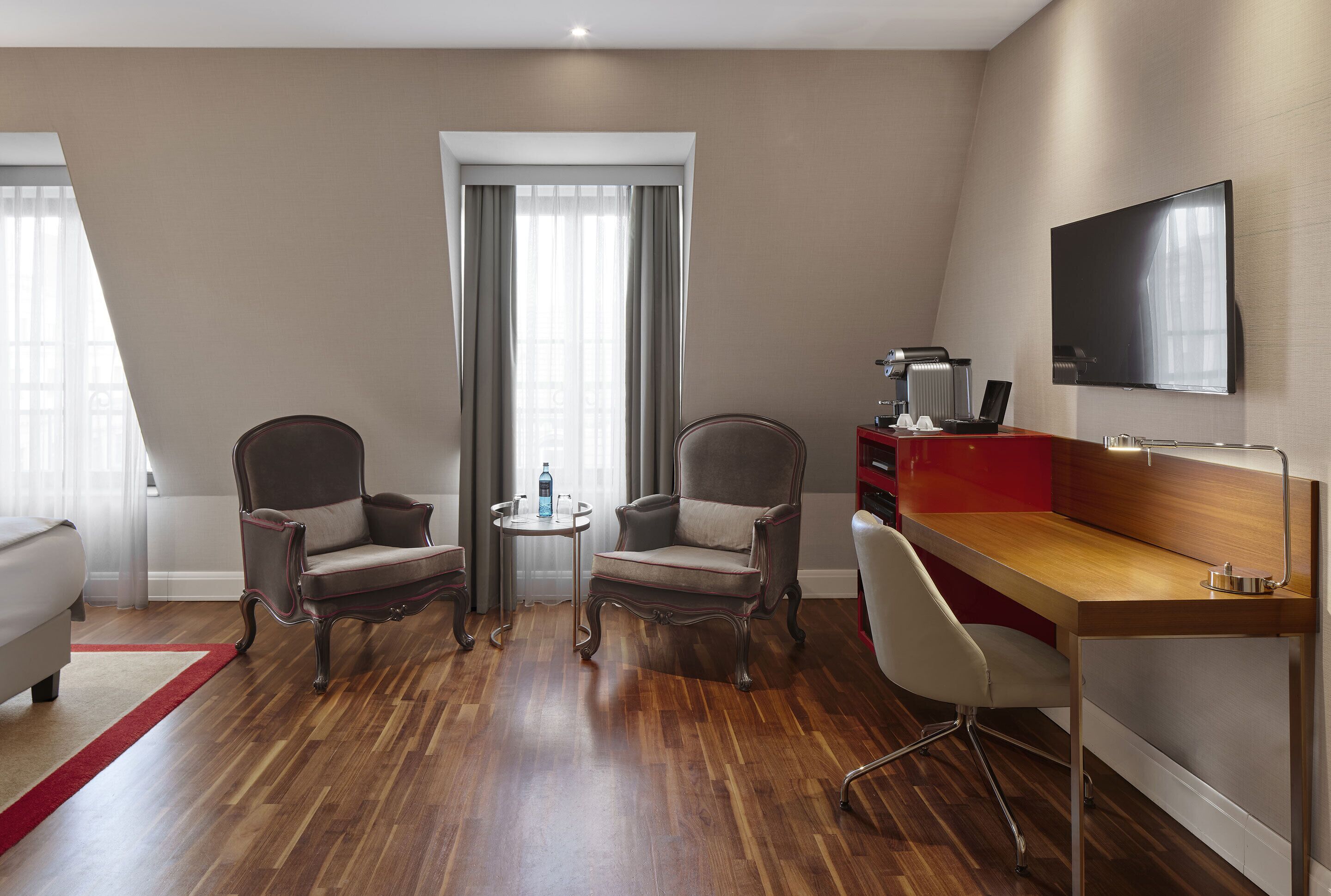 A hotel room with a wooden floor, two armchairs, a small round table, a desk with a chair, a TV mounted on the wall, and a window with curtains.