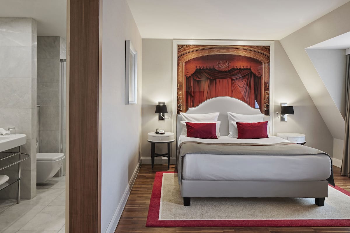 A modern bedroom with a large bed, a red accent wall, and a view of the city.