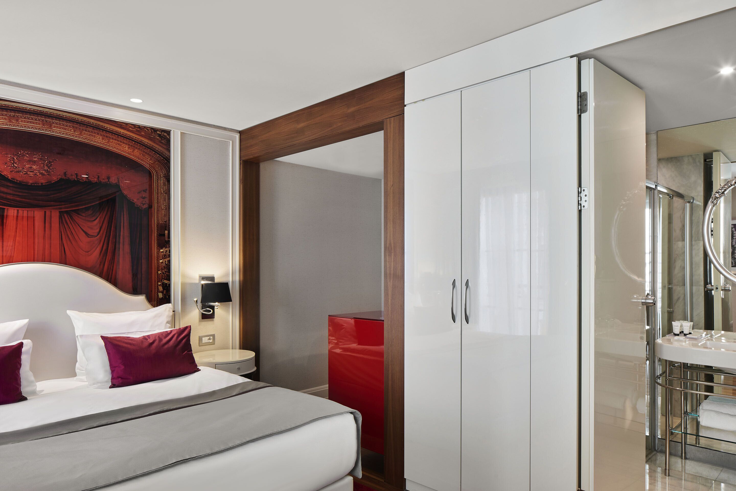 A modern hotel room with a large bed, a wardrobe, a red cabinet, and a bathroom with a mirror and sink.