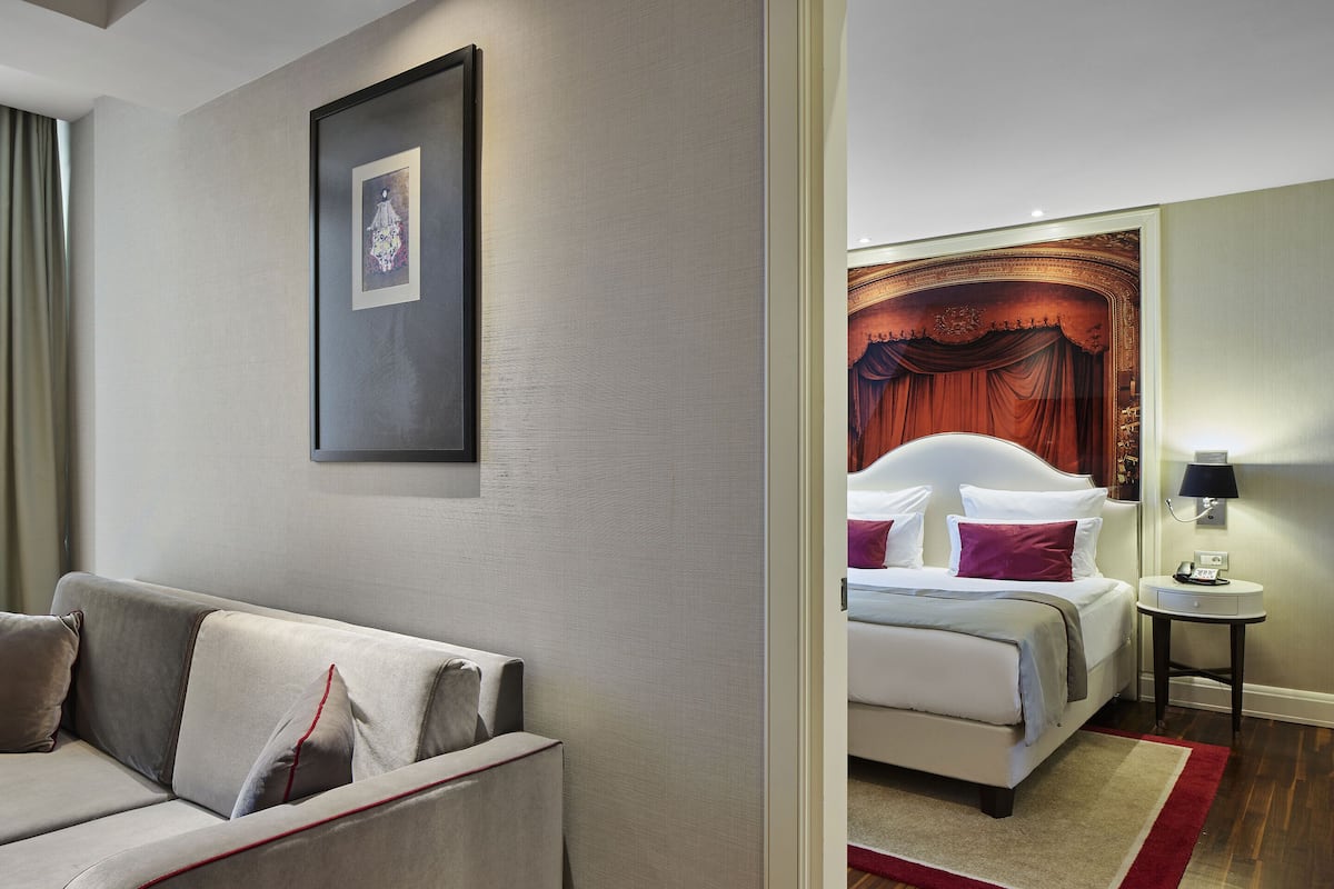 A hotel room with a sofa, a bed with white bedding and red pillows, a nightstand with a lamp, and a framed picture on the wall.