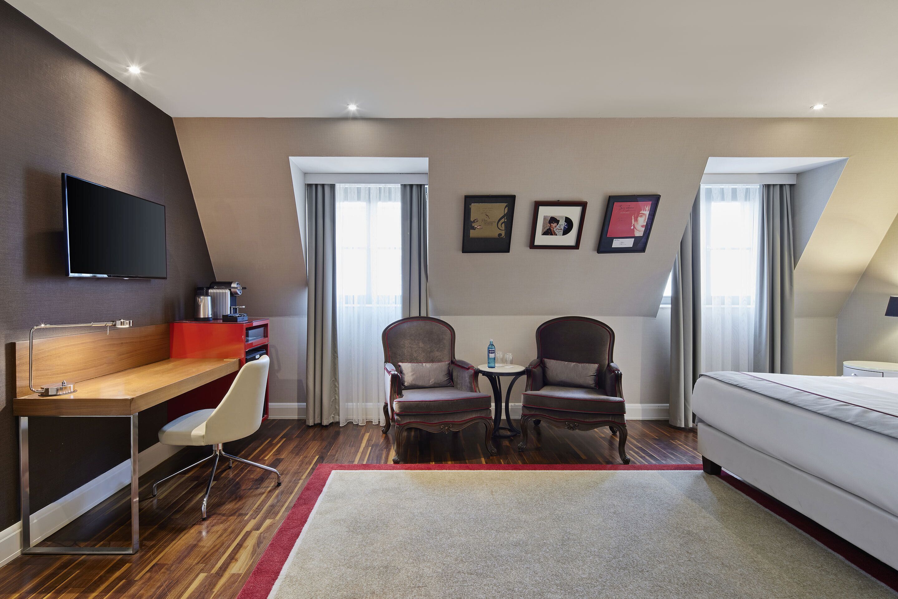 A hotel room with a desk, two armchairs, a bed, and a TV.