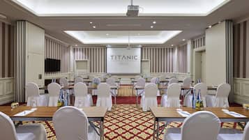 A conference room with a large screen displaying "TITANIC HOTELS" at the front. Rows of tables with chairs are set up for meetings.