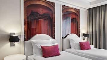 Quarto de hotel com duas camas, cada uma com roupa de cama branca e um travesseiro vermelho. Há duas luminárias de parede de cada lado das camas. O quarto tem um espelho grande com um pano de cortina vermelha decorativo ao fundo.