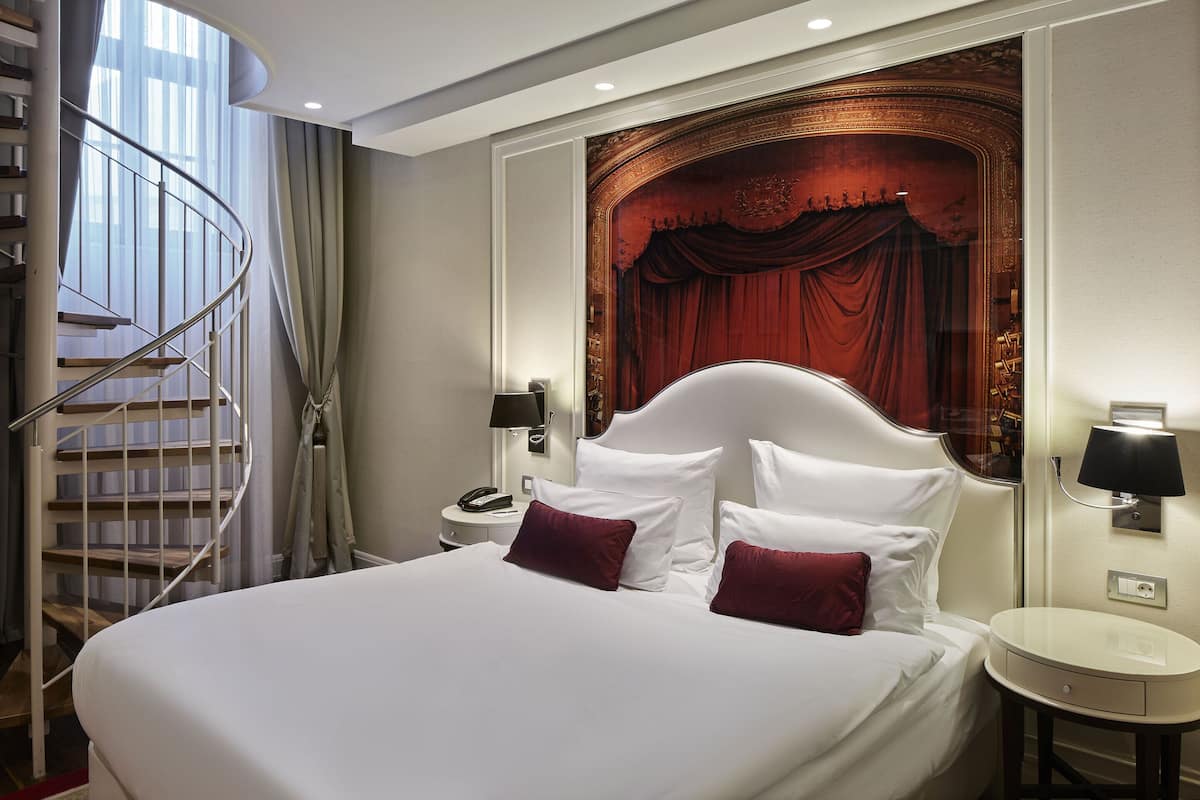 A hotel room with a large bed, a curved staircase, a round bedside table, and a wall-mounted lamp.