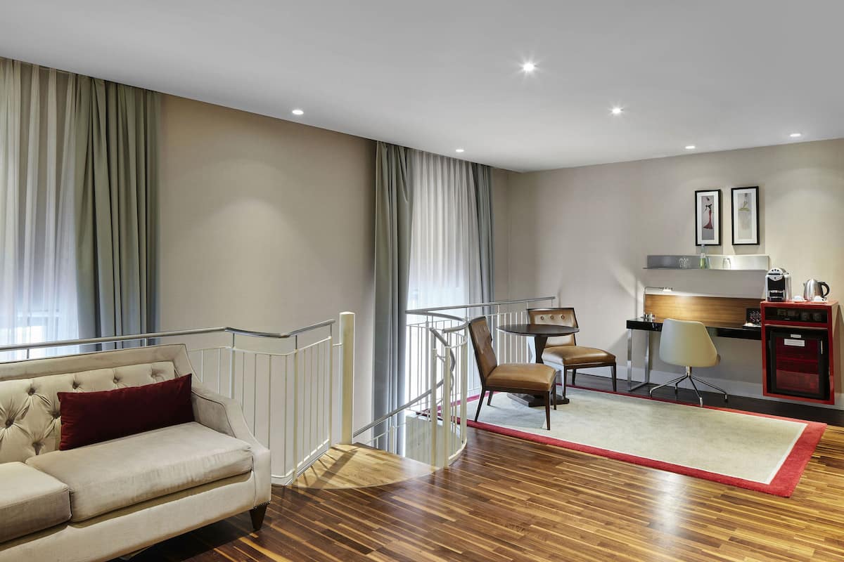 A modern hotel room with a sofa, a dining area, a kitchen, and a staircase.