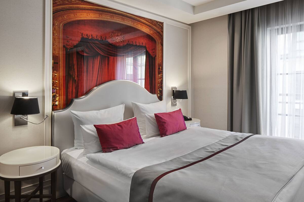 A neatly made bed with white and red pillows, a round bedside table, and a wall-mounted lamp.