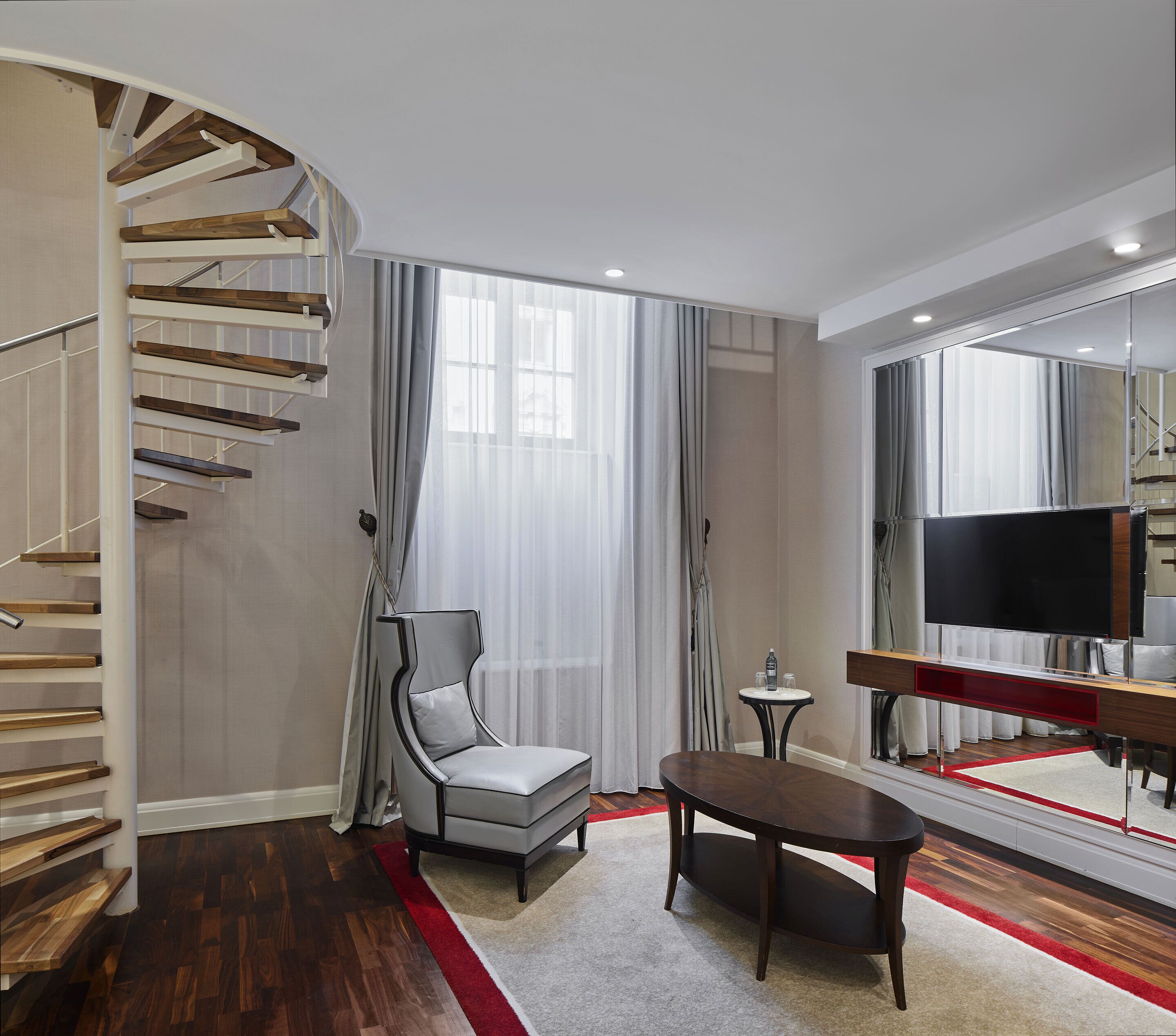 A modern living room with a curved staircase, a comfortable armchair, a small round table, and a flat-screen TV.
