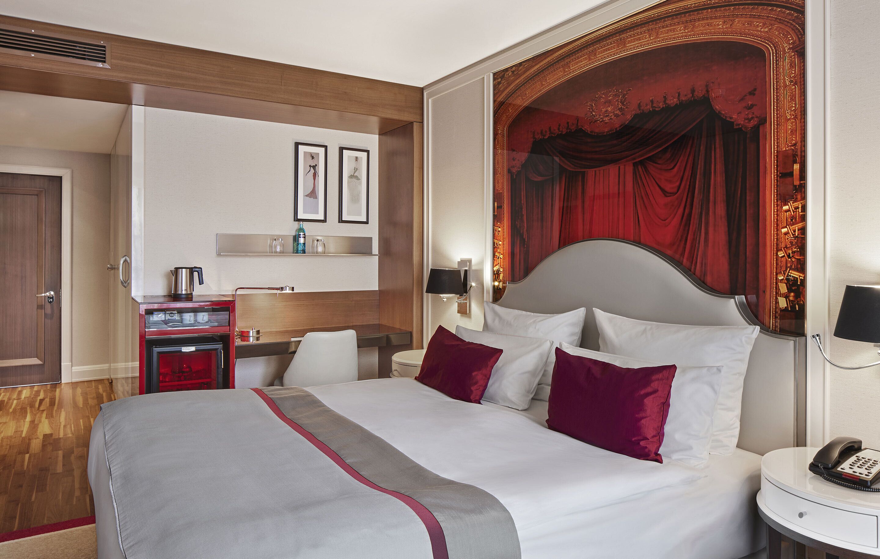 A hotel room with a large bed, a desk with a kettle, a minibar, and a red curtain backdrop.