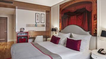 A hotel room with a large bed, a desk with a kettle, a minibar, and a red curtain backdrop.