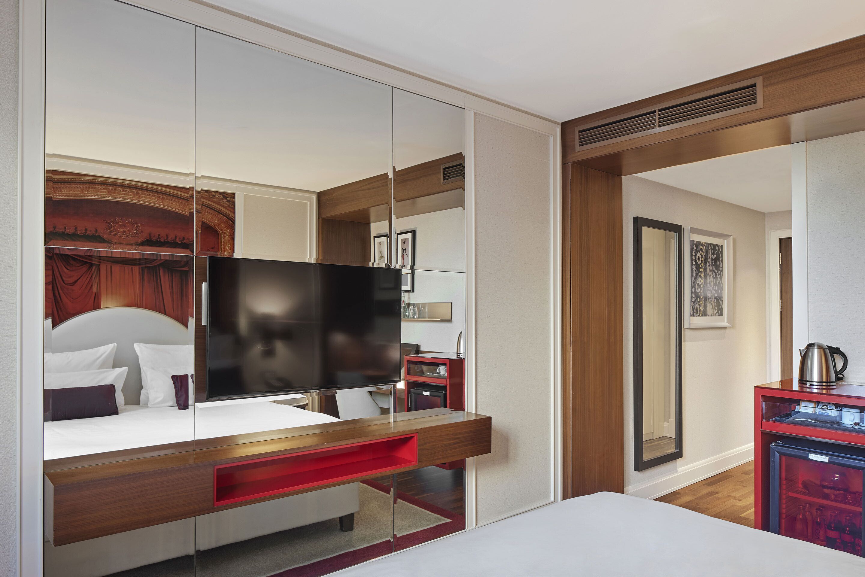 Una habitación de hotel moderna con una cama grande, un televisor, un minibar rojo y un espejo que refleja la decoración de la habitación.