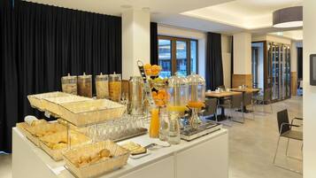Un bufé de desayuno con diversos alimentos y bebidas, una zona de estar con mesas y sillas, y vistas al exterior a través de grandes ventanales.
