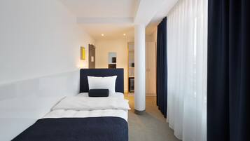 Une chambre d’hôtel avec un lit, une table de chevet, un bureau et une chaise. Il y a une fenêtre avec des rideaux et une vue sur le couloir.
