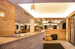Un'area di reception di un hotel con una scrivania in legno, un vaso con fiori, una lampada a sospensione moderna e un angolo salotto con una sedia e un tavolino.