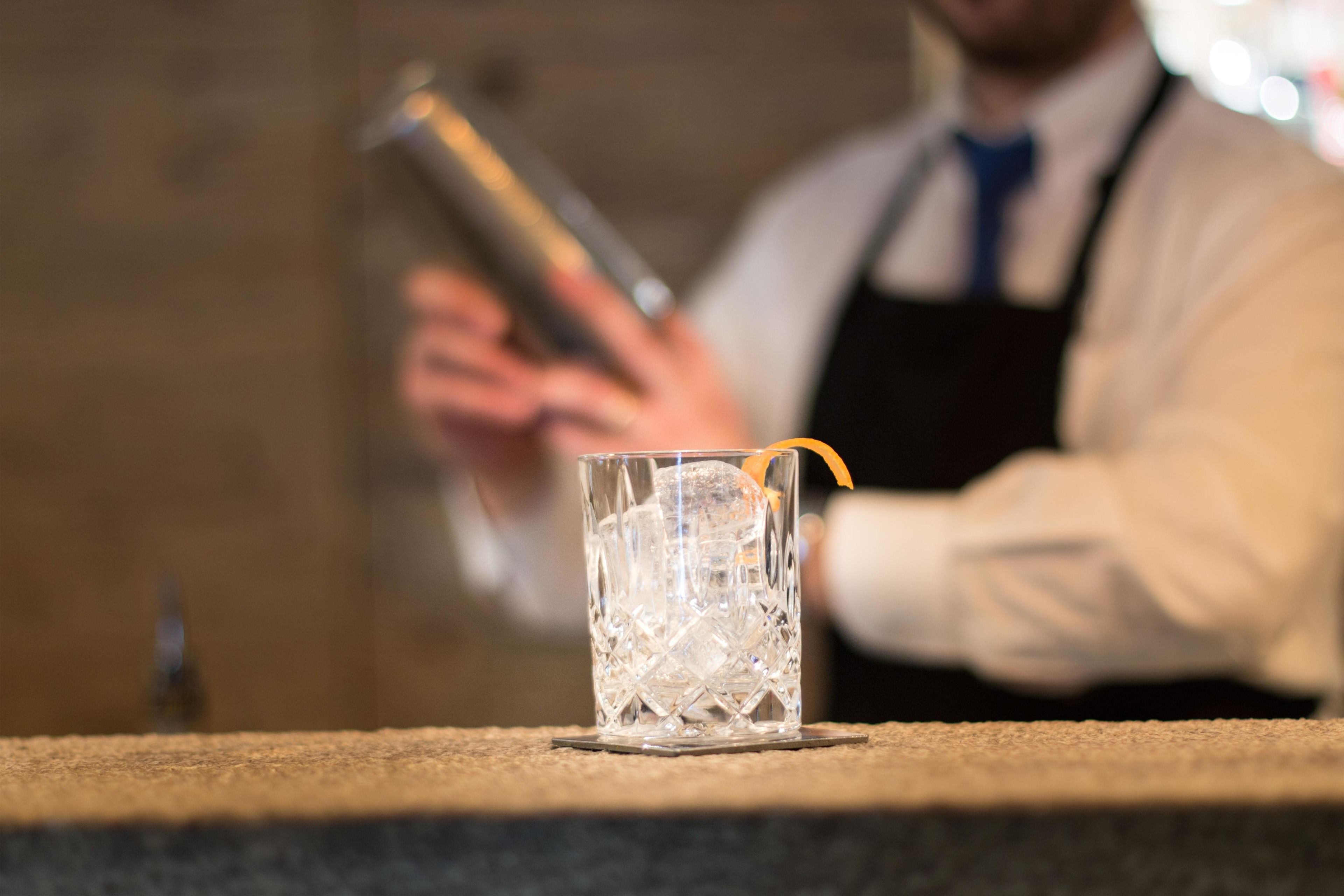Een barruimte met een helder glas gevuld met ijs en een cocktailgarnituur, en een barman die op de achtergrond een drankje bereidt.