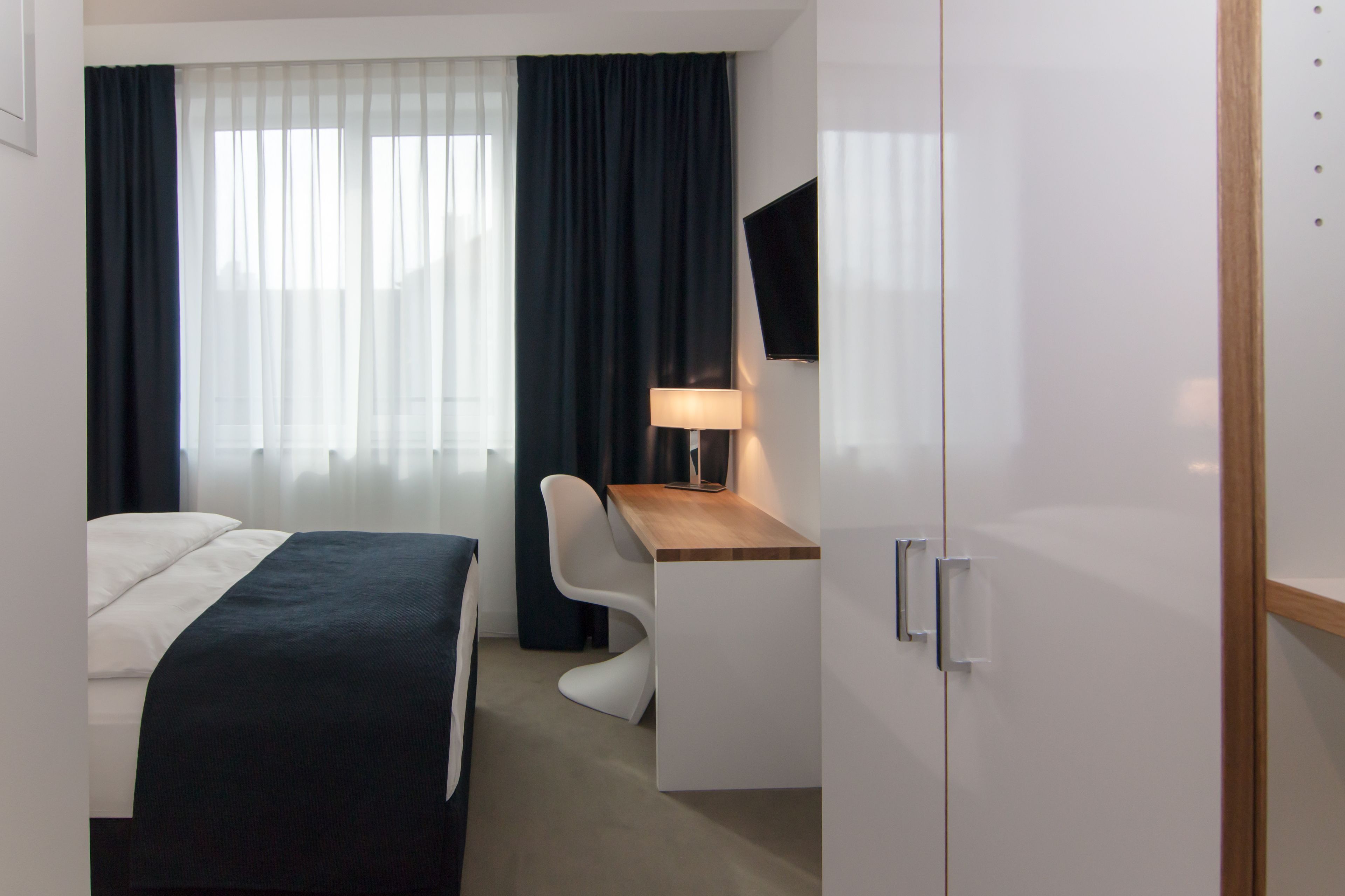 A hotel room with a bed, a desk, a chair, a TV, and a wardrobe.