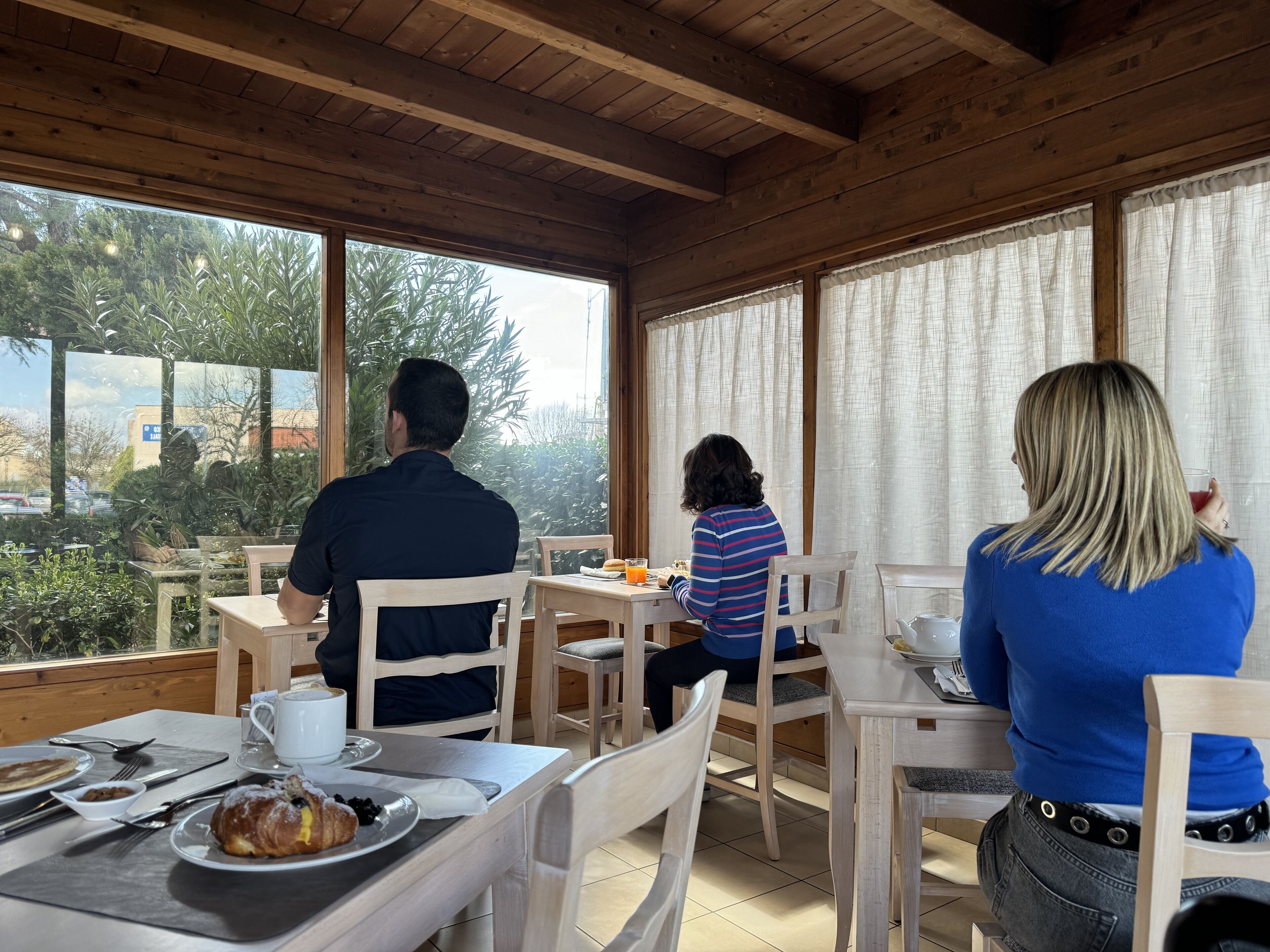 Ein Zimmer mit Holzwänden, Tischen und Stühlen, großen Fenstern mit Vorhängen und Blick ins Grüne.