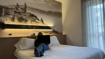 A hotel room with a bed, a nightstand, a lamp, and a large wall painting of a town.